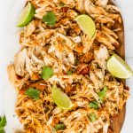 Shredded Mexican Chicken Garnished with Cilantro and sliced limes on cutting board on top of white background