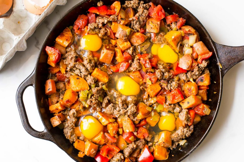 Sweet potato hash with sausage and five cracked raw eggs in cast iron skillet