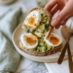 Hand holding a slice of avocado pesto toast with halved jammy eggs and thick orange yolks
