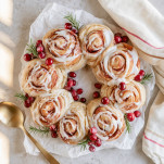 Glazed cinnamon roll wreath topped with cranberries and rosemary on parchment paper